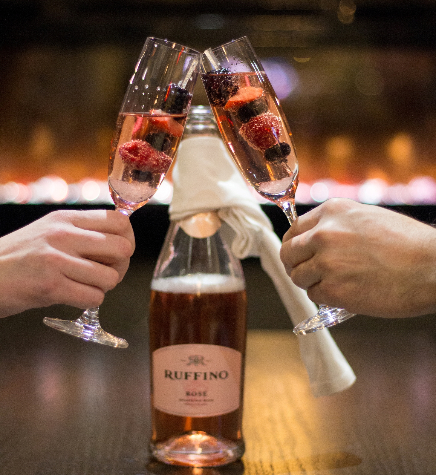 A picture of two people clinking their glasses together. The glass has Rose with strawberries, raspberries, blueberries and blackberries in it.